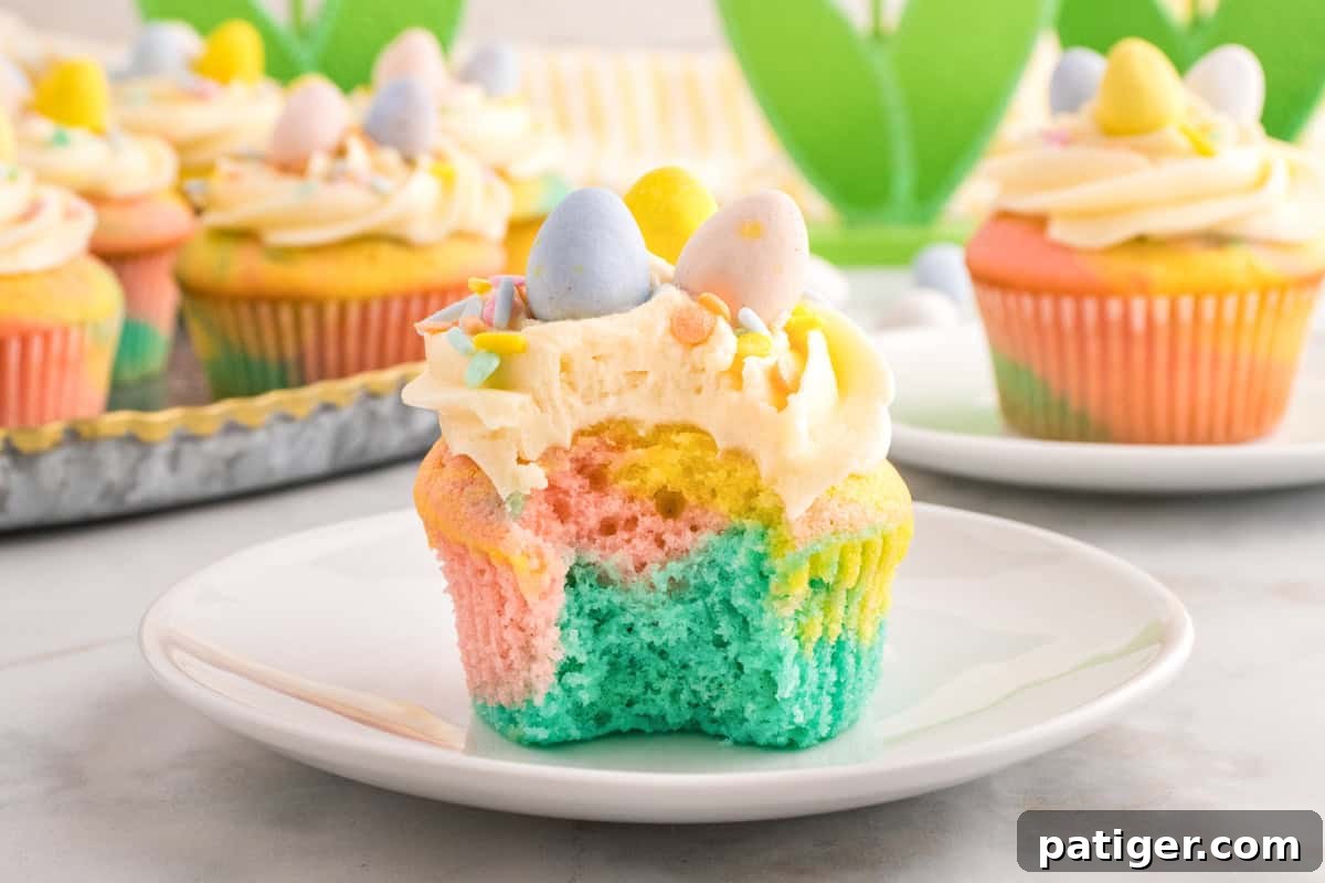 Hoppy Easter Cupcake Treats 3 A close-up of a pastel swirl Easter cupcake with a bite taken out, revealing soft layers of blue, pink, and yellow cake. The cupcake is topped with piped buttercream frosting, pastel sprinkles, and three mini chocolate eggs in blue, yellow, and pink.