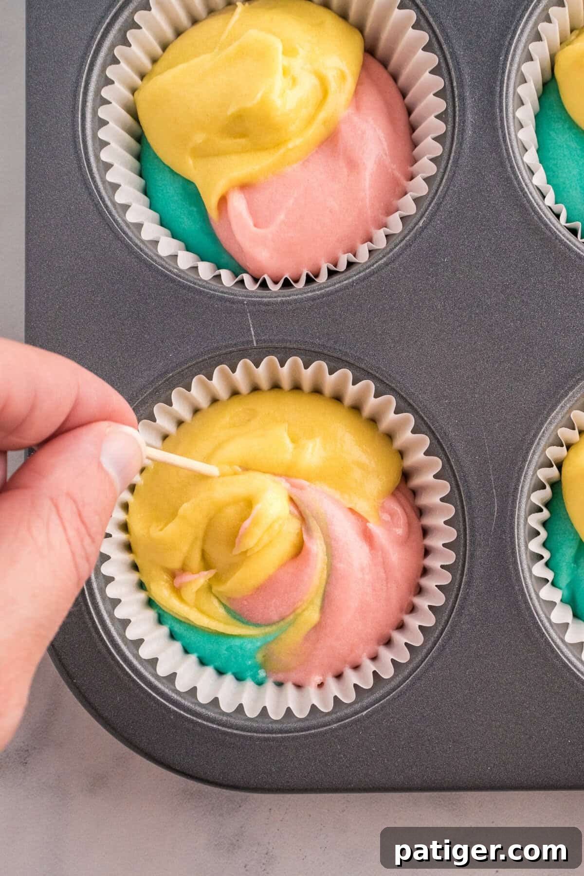 Hoppy Easter Cupcake Treats 7 A hand using a toothpick to swirl yellow, pink, and blue cupcake batter inside a paper liner placed in a muffin pan. The colors blend slightly, creating a marbled effect before baking.