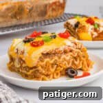 A slice of taco lasagna with noodles on a white plate, topped with melted cheese, olives, tomatoes, and green onions.