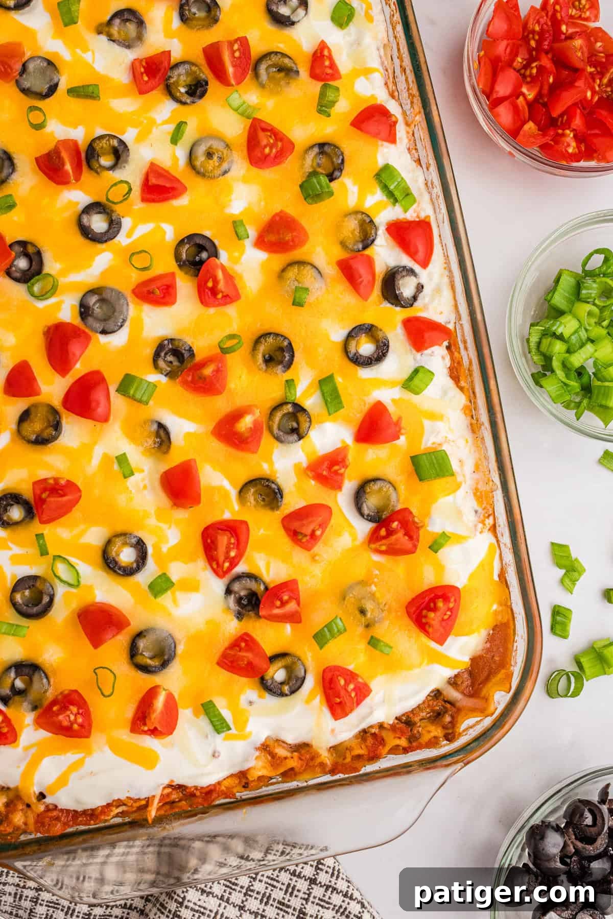 An inviting overhead view of a Mexican taco lasagna, generously topped with melted cheese, vibrant diced tomatoes, thinly sliced black olives, and fresh green onions, ready to be served.