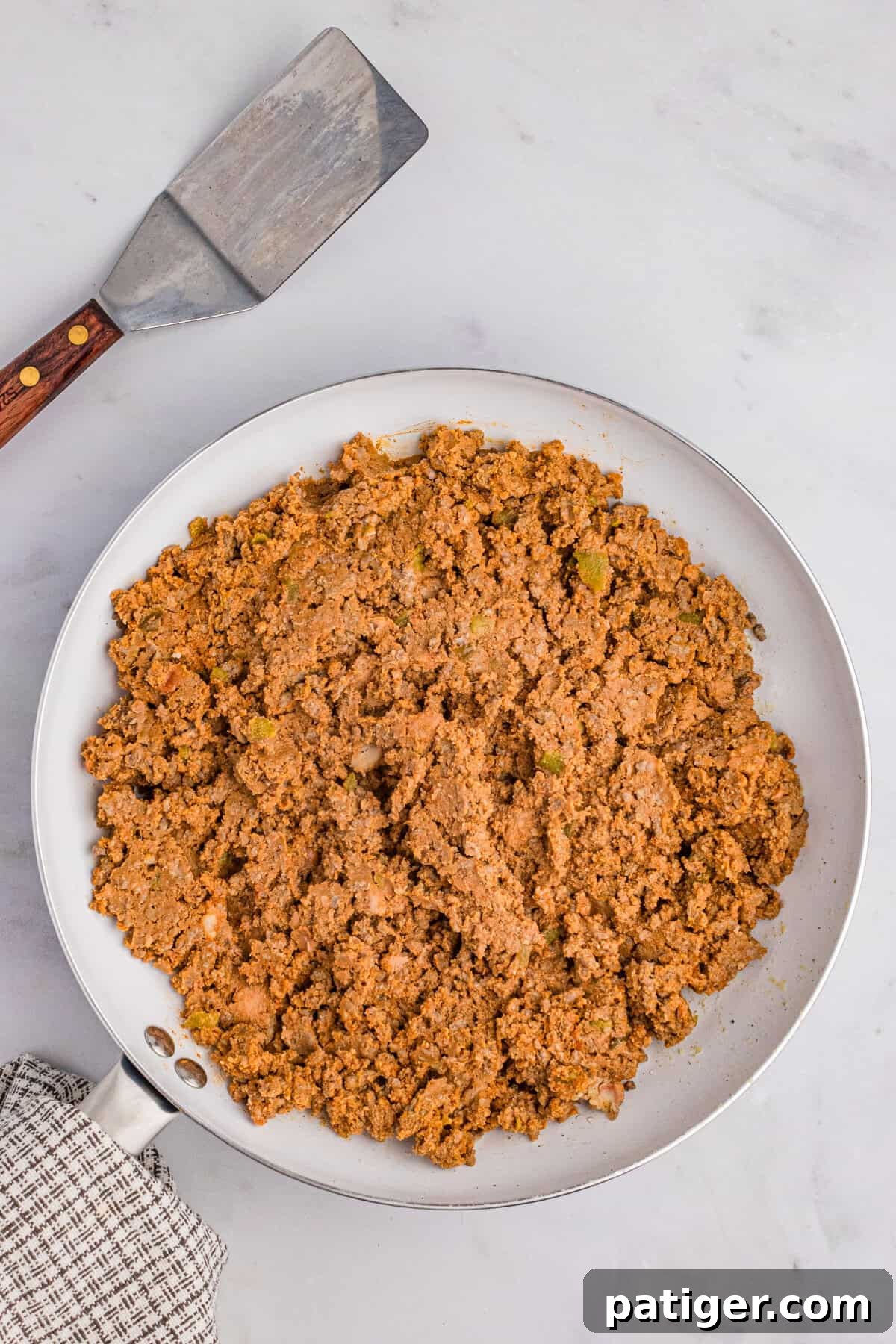 A large white skillet filled with perfectly seasoned taco meat, mixed with refried beans, ready for layering, with a spatula resting nearby.