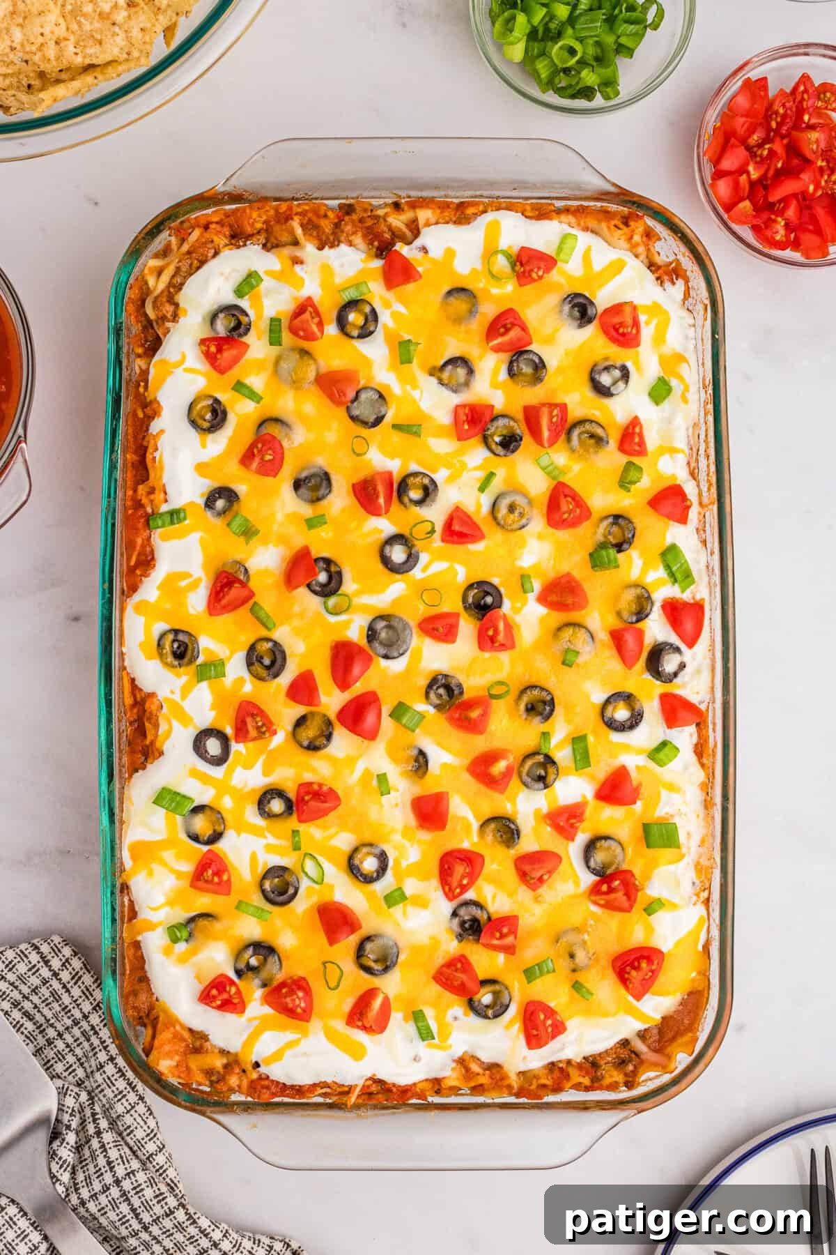A bountiful taco lasagna nestled in a glass 9x13 baking dish, lavishly topped with creamy sour cream, melted cheese, black olives, diced tomatoes, and vibrant green onions, ready to be enjoyed.
