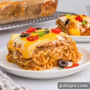 A slice of taco lasagna with noodles on a white plate, topped with melted cheese, olives, tomatoes, and green onions.