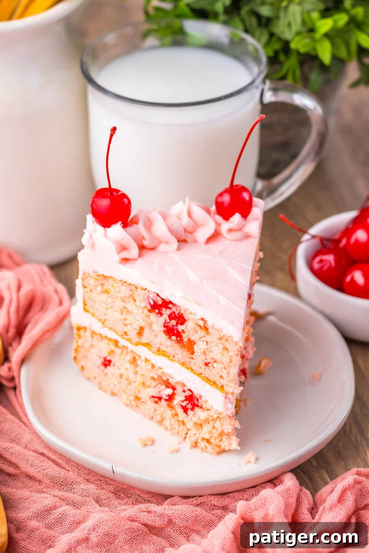 A beautifully sliced cherry chip cake, revealing two moist layers of light pink cake speckled generously with bits of red maraschino cherries. The layers are filled and topped with a creamy pink frosting, adorned with elegant swirls and whole, glistening cherries.