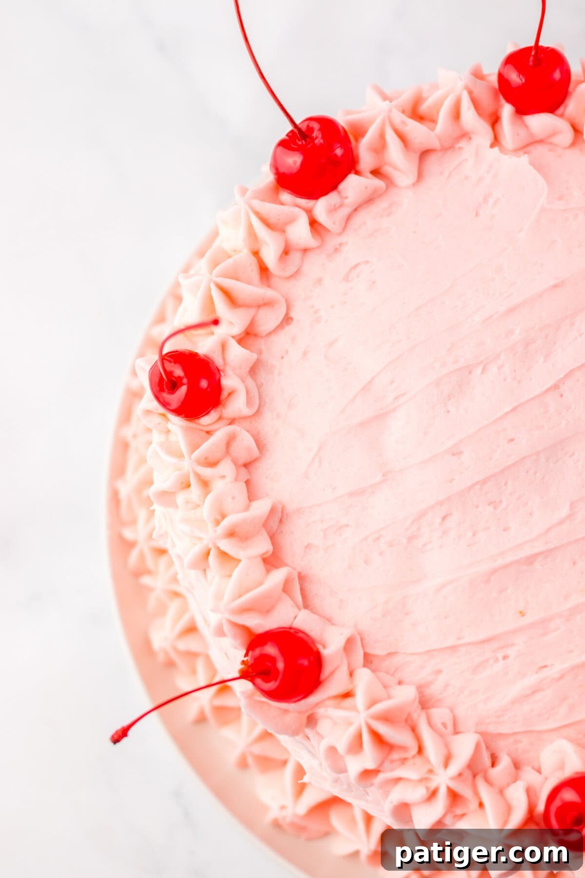 An inviting overhead shot of a perfectly round maraschino cherry cake, adorned with smooth pink buttercream frosting. The edges are beautifully decorated with intricately piped rosettes, and whole, bright red maraschino cherries are elegantly spaced on top for a finishing touch.