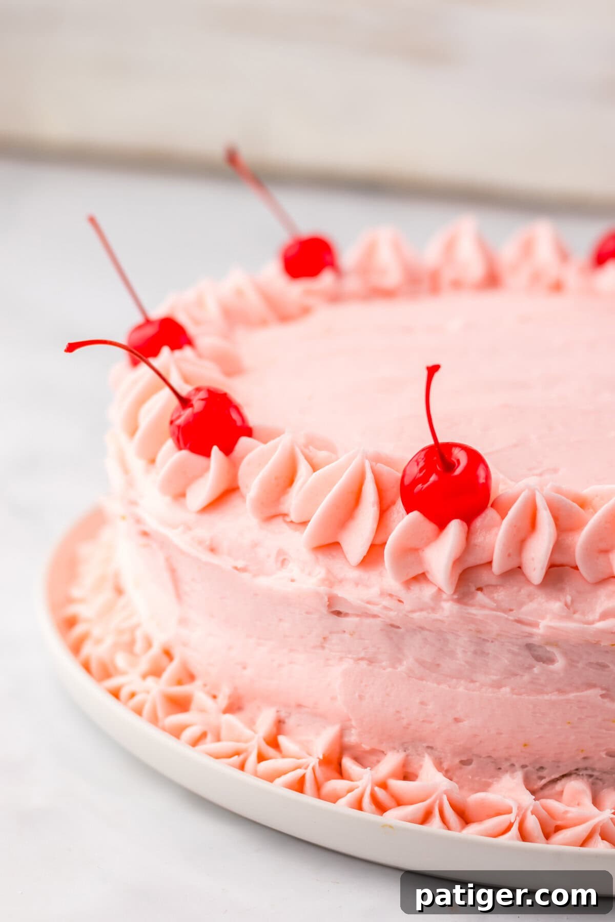 A visually appealing maraschino cherry cake, elegantly frosted with pink buttercream and decorated with charming piped rosettes around the top edge, featuring glistening whole maraschino cherries as a vibrant final flourish.
