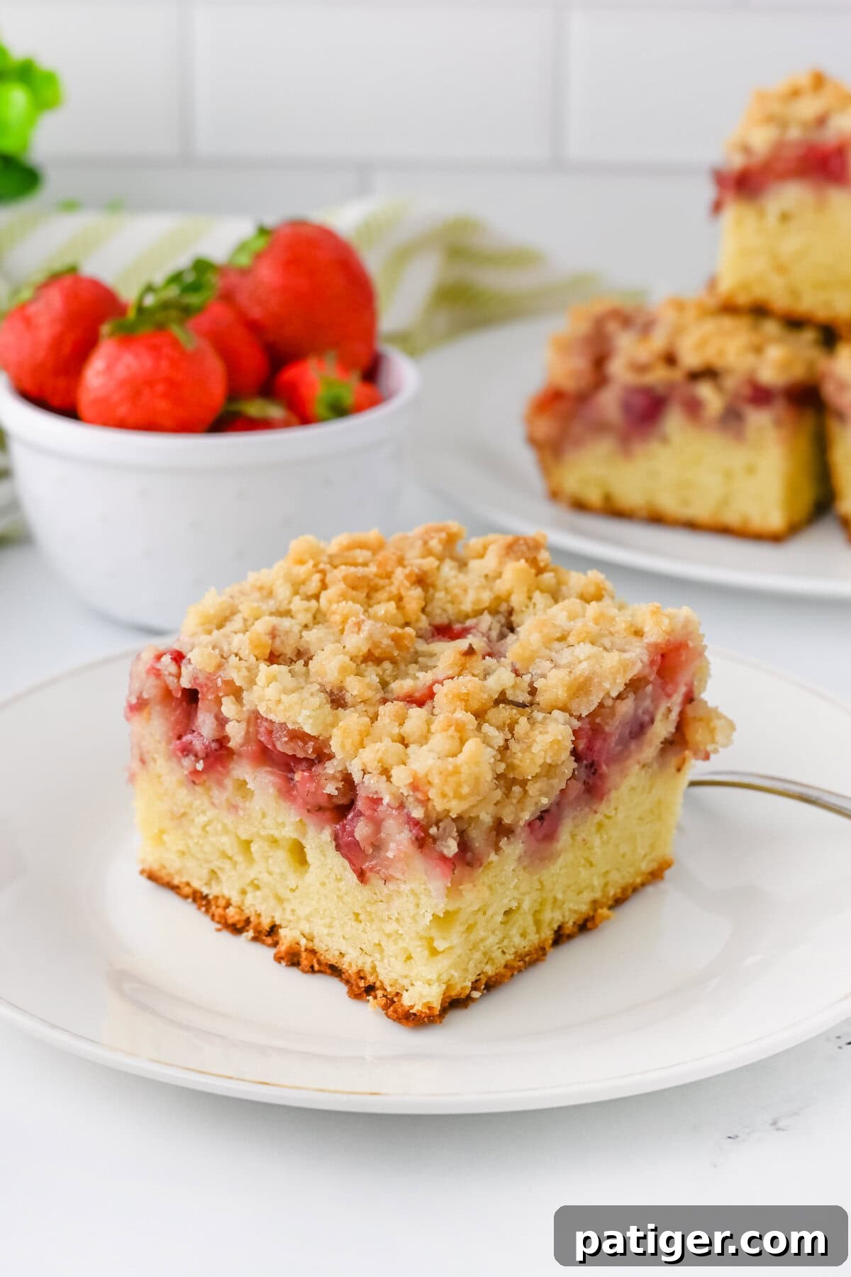 Scrumptious Strawberry Crumb Cake 2 A beautifully presented slice of strawberry crumb cake, showcasing its moist vanilla cake base, a vibrant layer of baked strawberries, and a generous golden crumb topping. In the background, a rustic bowl overflows with fresh, ripe strawberries, alongside additional slices of the tempting cake, ready to be enjoyed.