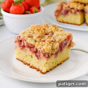 A slice of strawberry crumb cake with a moist vanilla cake base, a layer of baked strawberries, and a golden crumb topping. In the background, there is a bowl of fresh strawberries and additional cake slices.