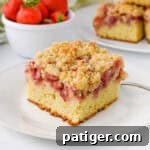 A slice of strawberry crumb cake with a moist vanilla cake base, a layer of baked strawberries, and a golden crumb topping. In the background, there is a bowl of fresh strawberries and additional cake slices.