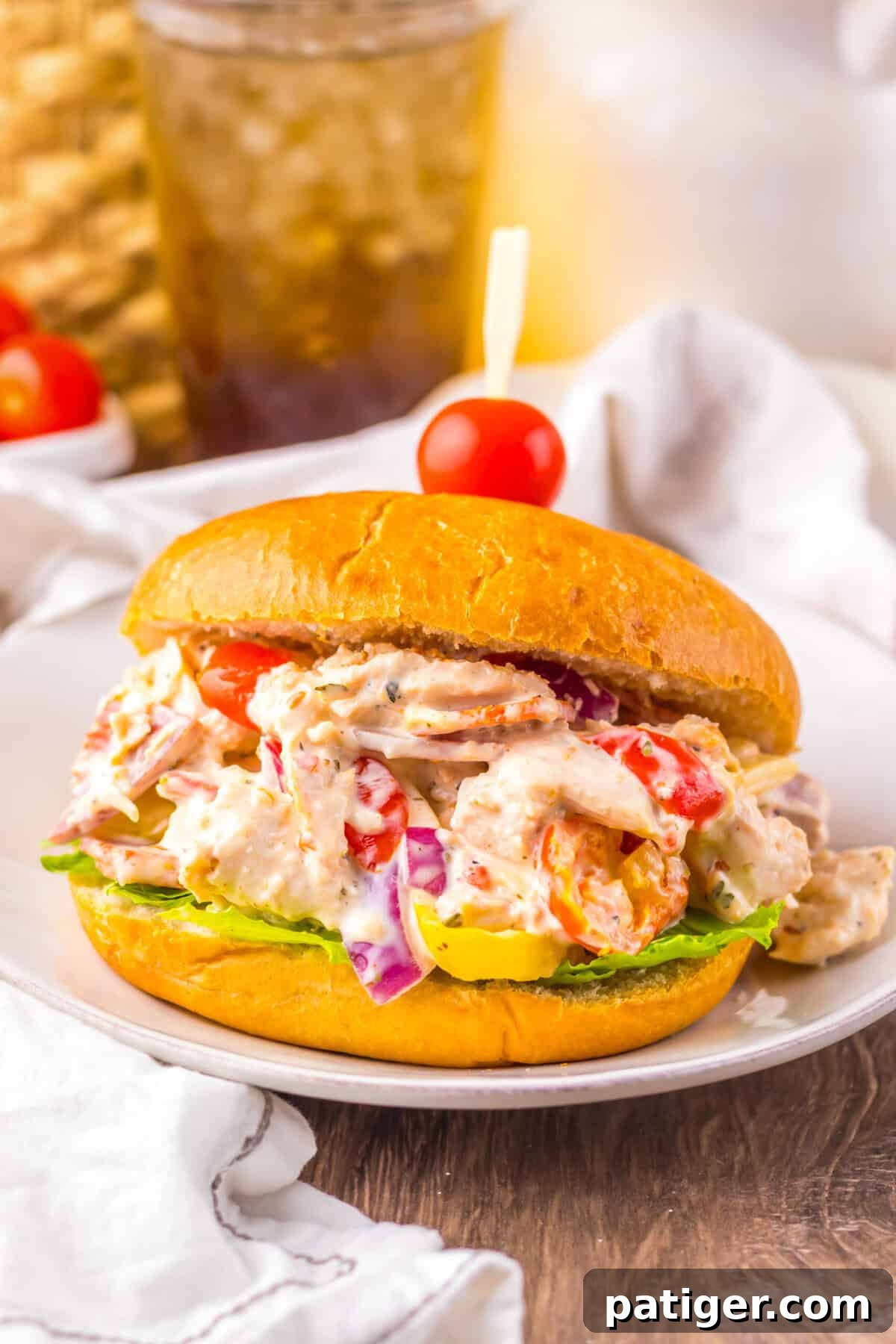 A sandwich filled with creamy Italian grinder chicken salad featuring shredded chicken, pepperoni, salami, provolone, cherry tomatoes, red onions, banana peppers, and lettuce on a golden bun, secured with a toothpick topped with a cherry tomato. 
