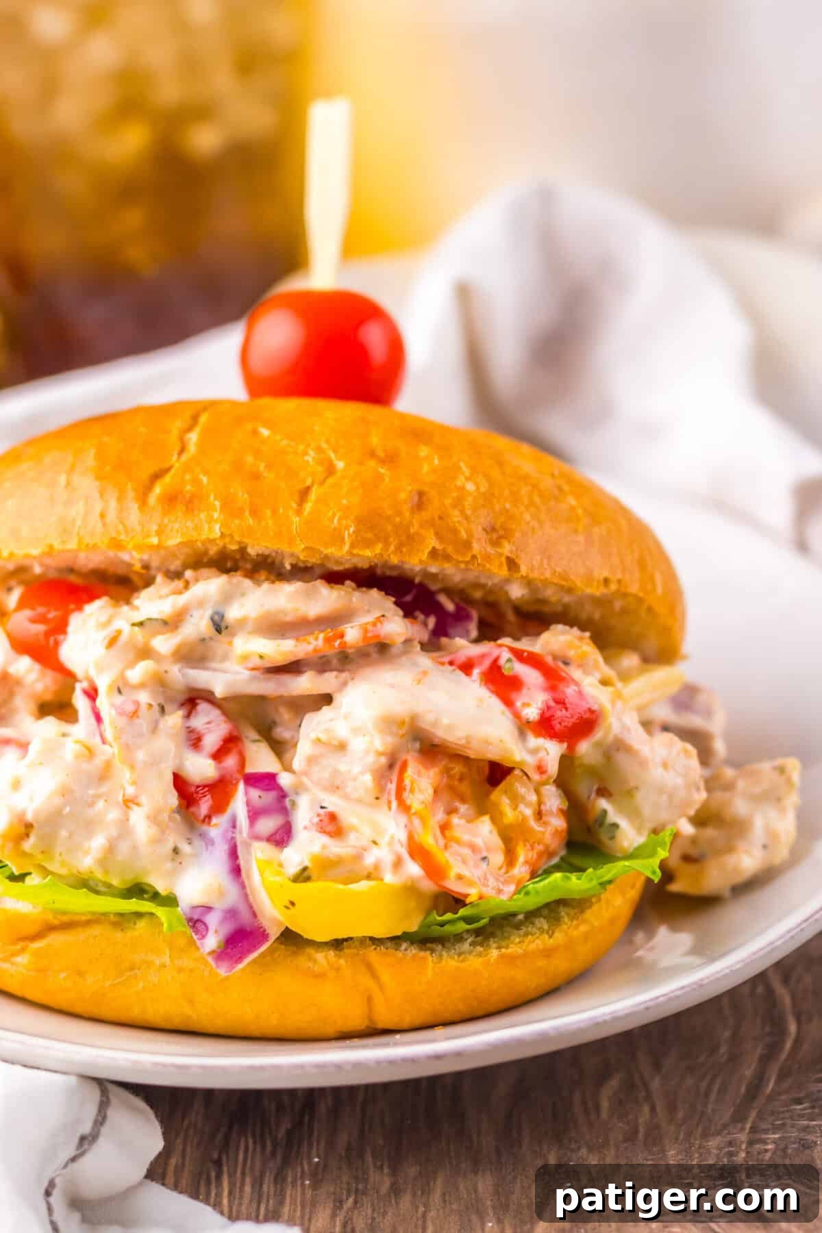 A close-up view of a sandwich stuffed with creamy chicken salad, showing visible chunks of chicken, red onion, cherry tomatoes, and banana peppers, with a cherry tomato on top of the bun.
