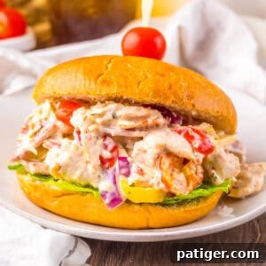 A sandwich filled with creamy Italian grinder chicken salad featuring shredded chicken, pepperoni, salami, provolone, cherry tomatoes, red onions, banana peppers, and lettuce on a golden bun, secured with a toothpick topped with a cherry tomato.