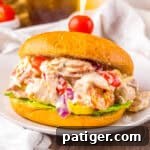 A sandwich filled with creamy Italian grinder chicken salad featuring shredded chicken, pepperoni, salami, provolone, cherry tomatoes, red onions, banana peppers, and lettuce on a golden bun, secured with a toothpick topped with a cherry tomato.