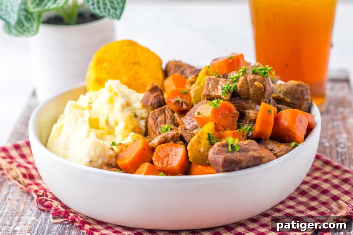 Side view of a bowl of crockpot pot roast, featuring stew beef, sliced carrots, celery, mashed potatoes, and a biscuit. The rich gravy glistens and the comfort food presentation is inviting.