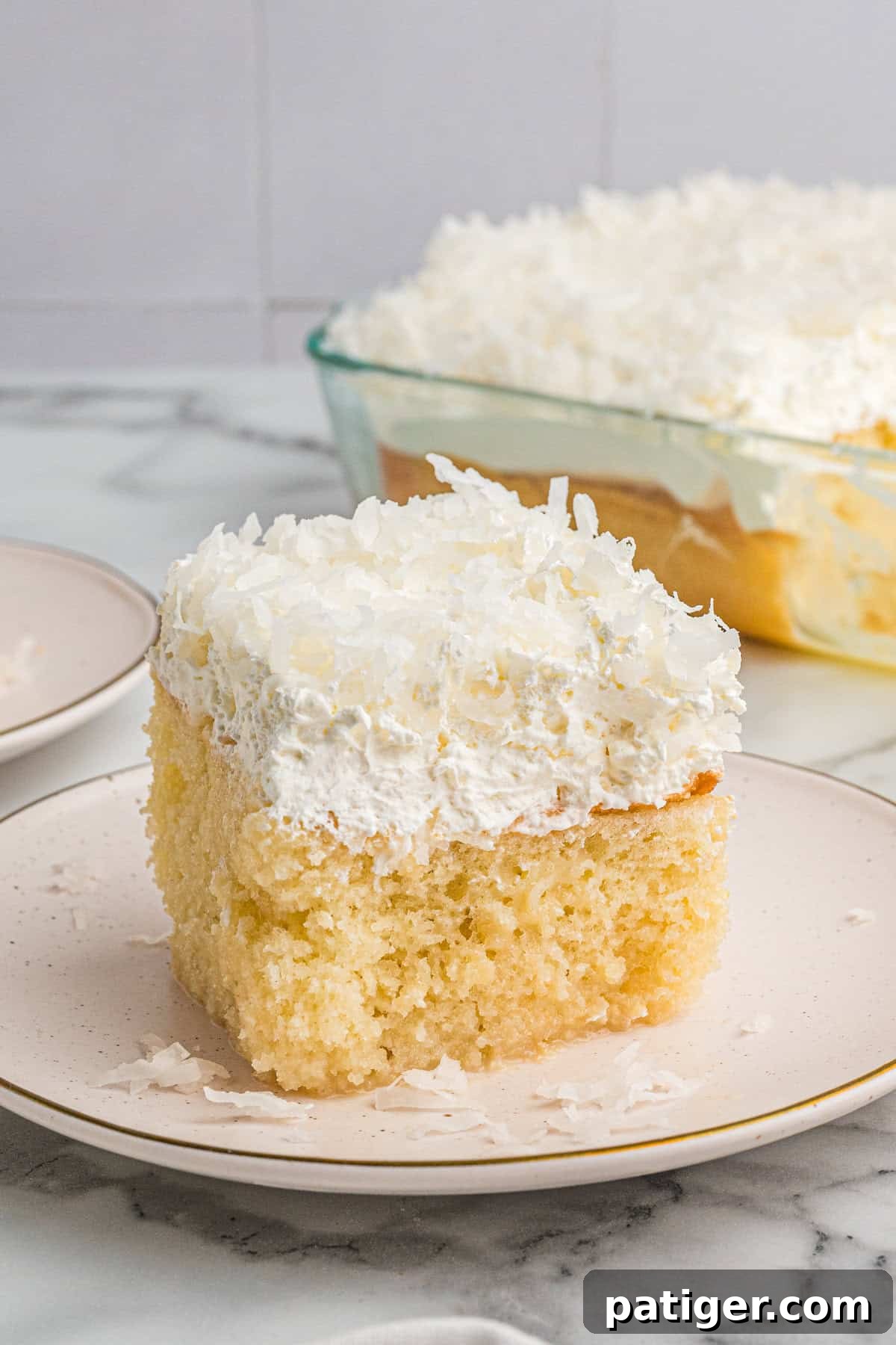 Tropical Coconut Poke Cake 2 A tall slice of coconut poke cake on a plate, showing its fluffy base, thick whipped topping, and coconut sprinkled on top.