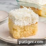 A square slice of coconut poke cake on a white plate with a fluffy whipped topping and shredded coconut. The full cake in a baking dish is visible in the background.