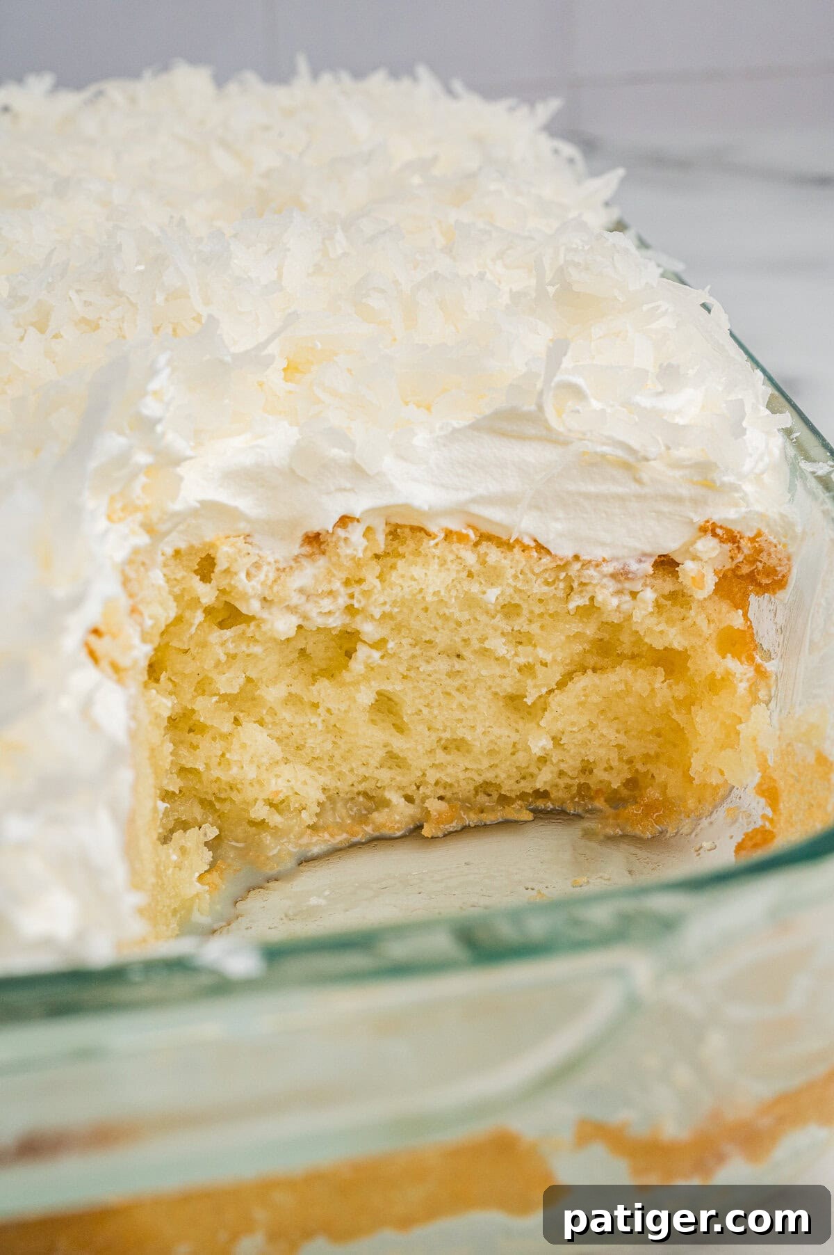 Tropical Coconut Poke Cake 7 A close-up view of a moist yellow coconut poke cake with a square slice removed, revealing its soft texture. The cake is topped with fluffy whipped topping and shredded coconut in a glass baking dish.
