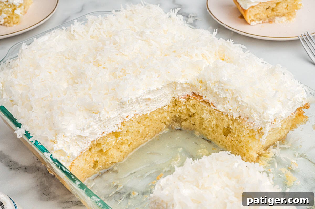 Tropical Coconut Poke Cake 9 A baked coconut poke cake in a glass dish with several slices missing. The cake has a fluffy white whipped topping and is generously sprinkled with shredded coconut.