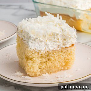 A square slice of coconut poke cake on a white plate with a fluffy whipped topping and shredded coconut. The full cake in a baking dish is visible in the background.