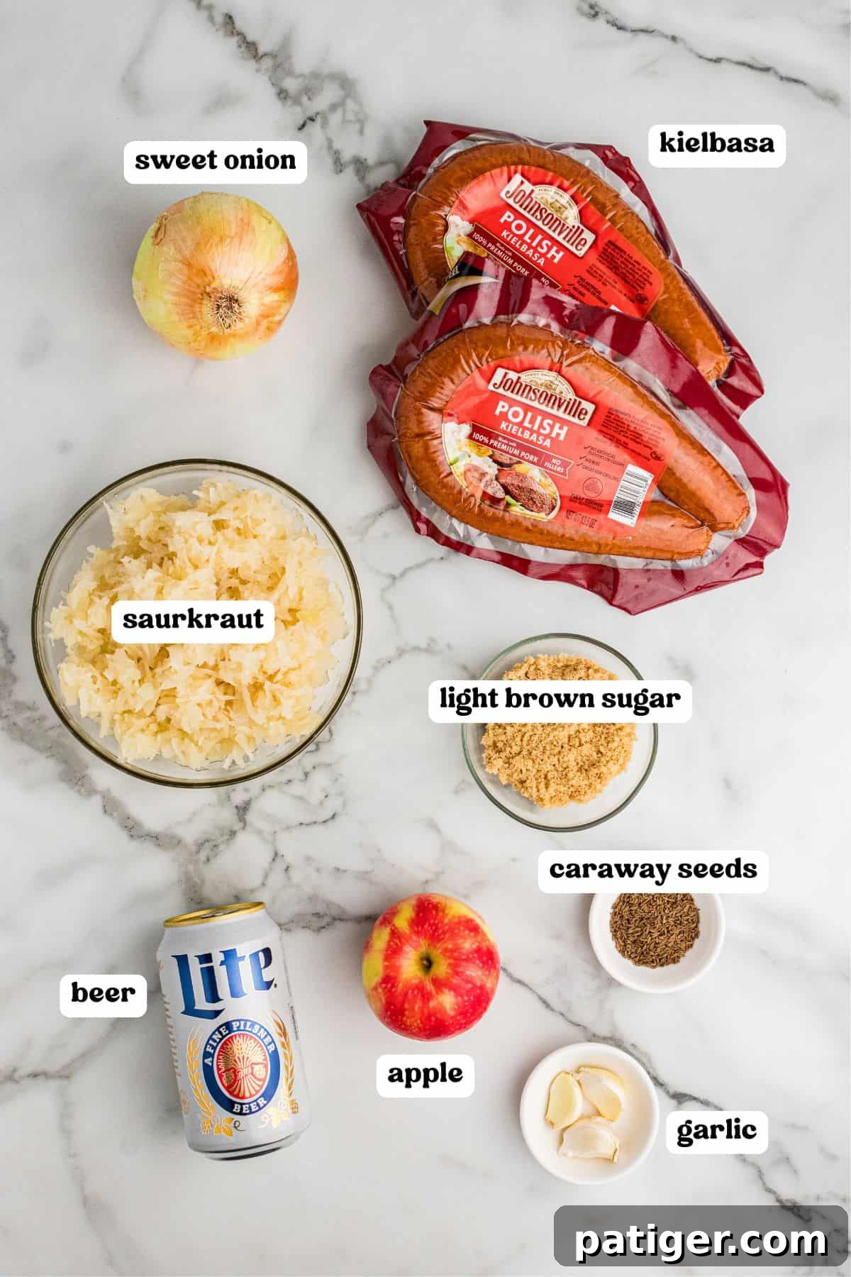Ingredients on a white marble surface: sweet onion, sauerkraut, light brown sugar, caraway seeds, garlic cloves, an apple, a can of Lite beer, and two packages of Johnsonville Polish kielbasa.