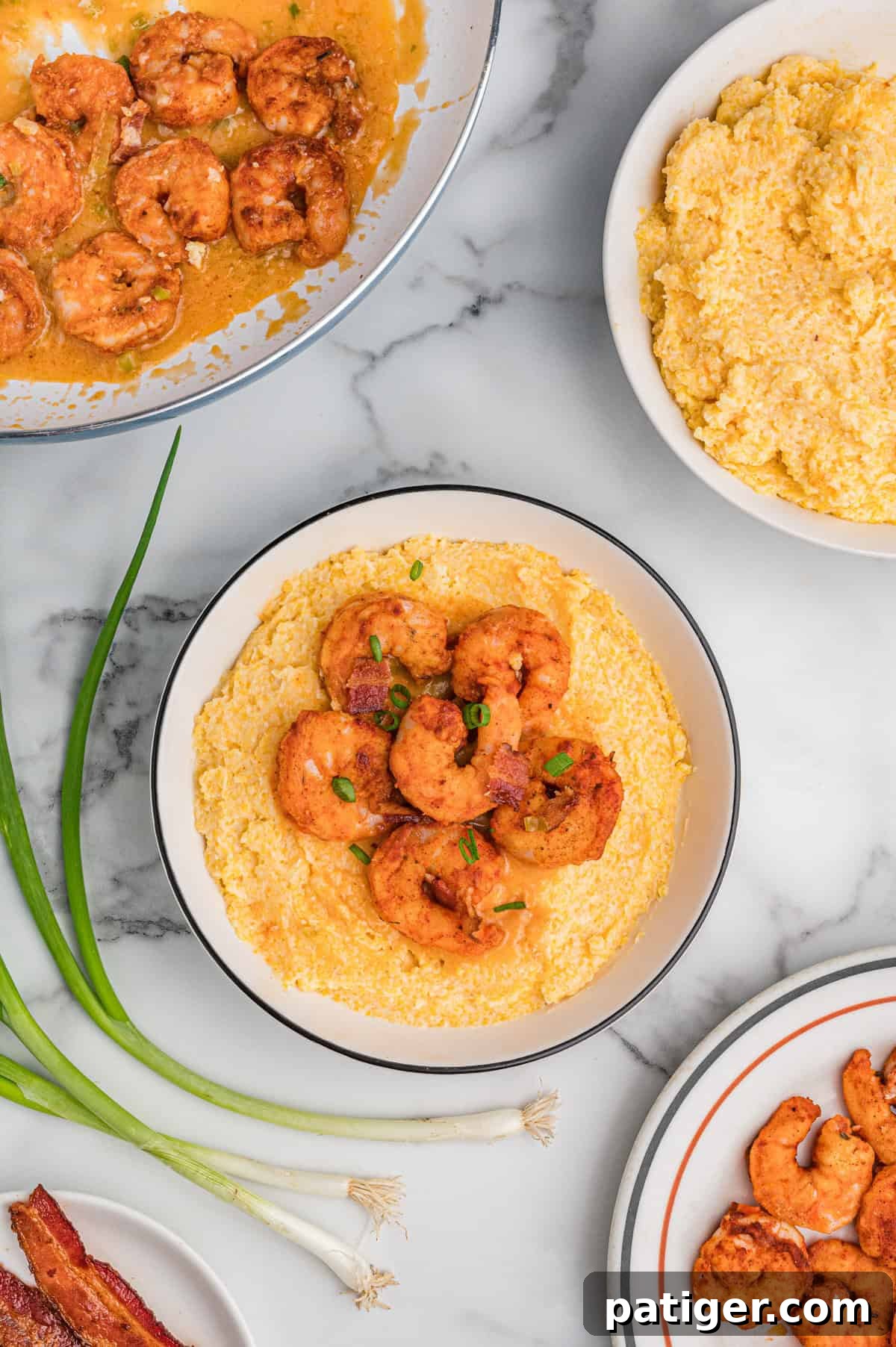 Loaded Cheesy Shrimp Grits 2 Overhead view of a bowl of cheesy shrimp and grits with bacon and green onions. Surrounding it are a skillet with saucy shrimp, a bowl of cheesy grits, a plate of cooked shrimp, fresh green onions, and a plate of bacon, all arranged on a marble surface.