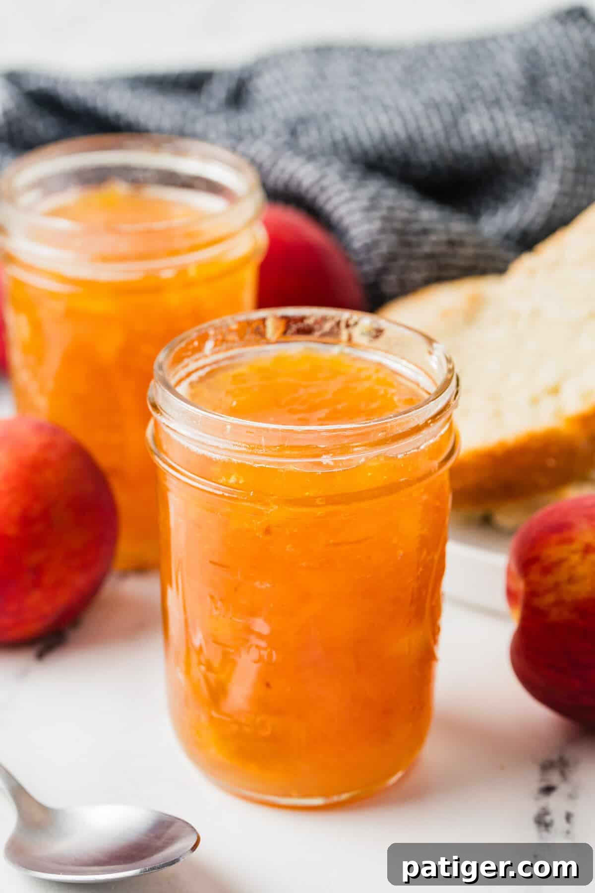 Two jars of homemade peach jam with whole peaches, a spoon, and a plate with sliced bread in the background.