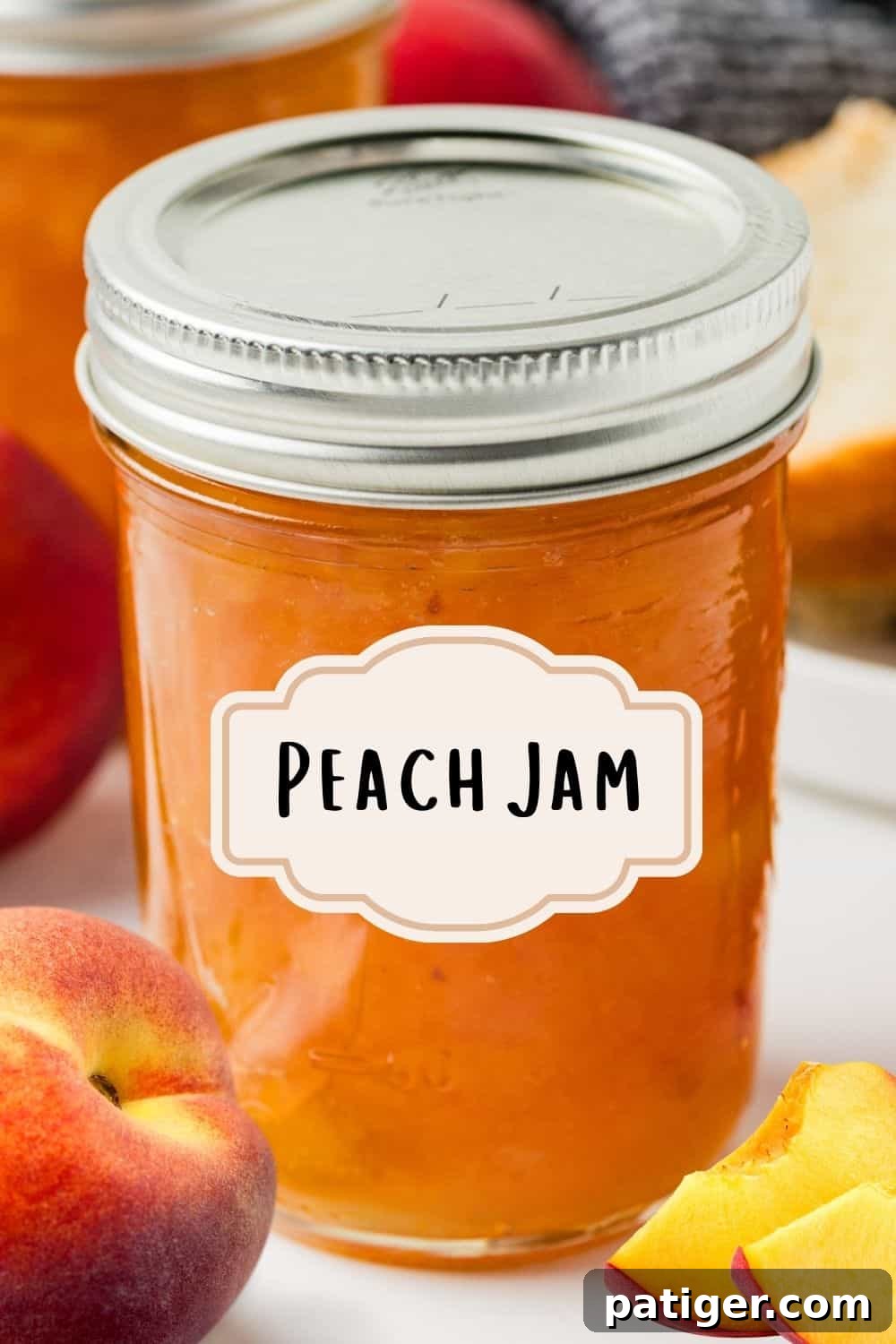 A sealed mason jar labeled “Peach Jam” sits on a marble surface surrounded by fresh peaches, a slice of bread, and another jar in the background.