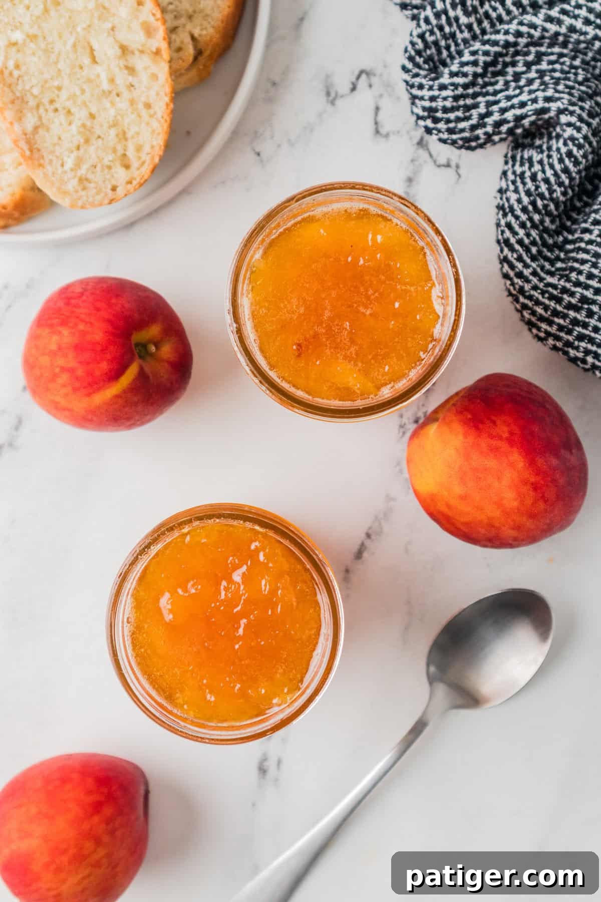 Overhead view showing two open jars of peach jam on a marble surface, surrounded by whole fresh peaches, a silver spoon, a black-and-white cloth, and a plate with slices of bread.