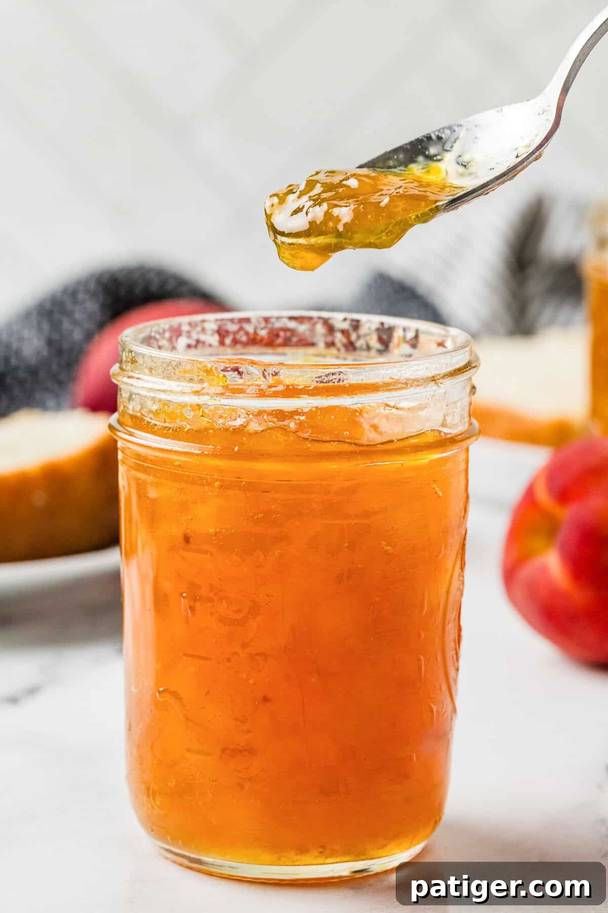 Close-up of a spoon lifting thick peach jam from an open mason jar, with a peach and bread in the background.