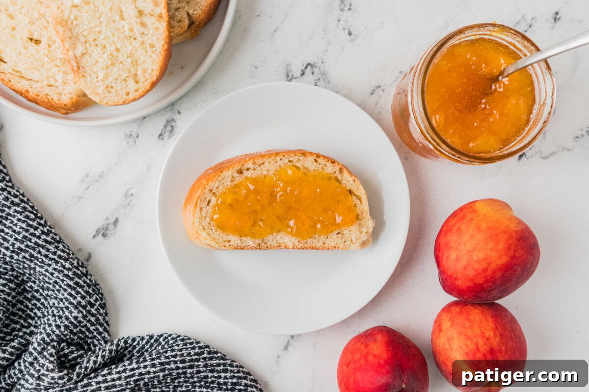 Slice of bread with peach jam on a plate next to a jar of homemade peach jam and peaches.
