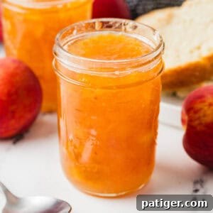 Jars of homemade peach jam with whole peaches, a spoon, and a plate with sliced bread in the background.