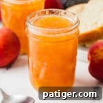 Jars of homemade peach jam with whole peaches, a spoon, and a plate with sliced bread in the background.