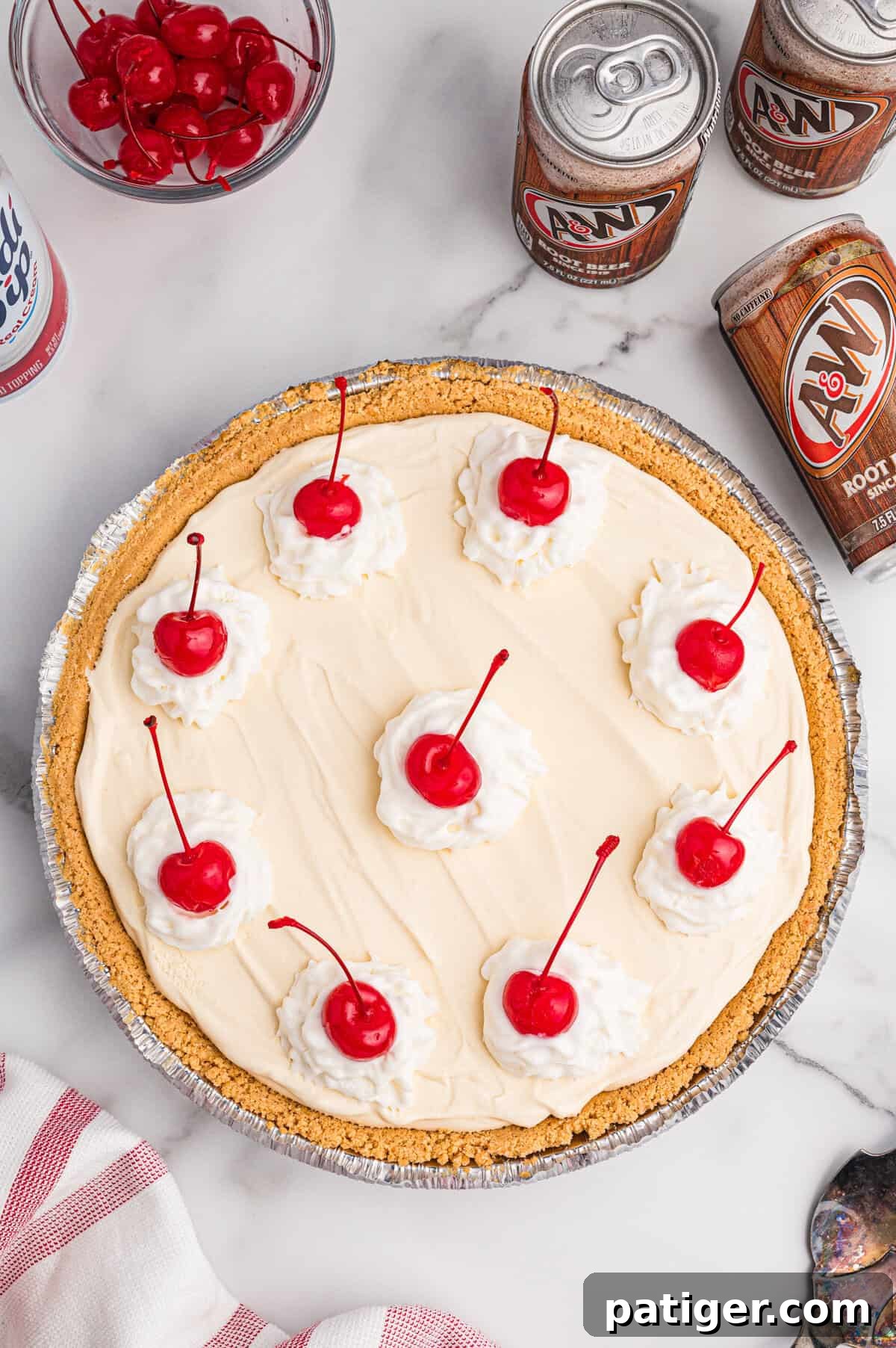 Overhead view of a full root beer float pie with whipped cream dollops and cherries; surrounded by root beer cans and a bowl of cherries.