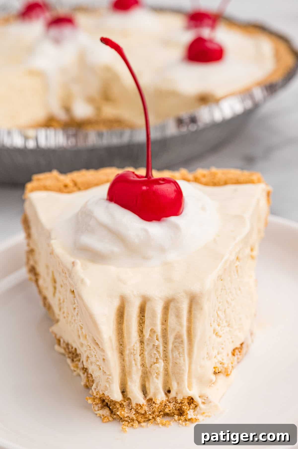 A close-up of a plated slice of root beer float pie with a graham cracker crust. The pie slice has creamy, slightly melted filling with visible fork marks and is topped with whipped cream and a maraschino cherry. 