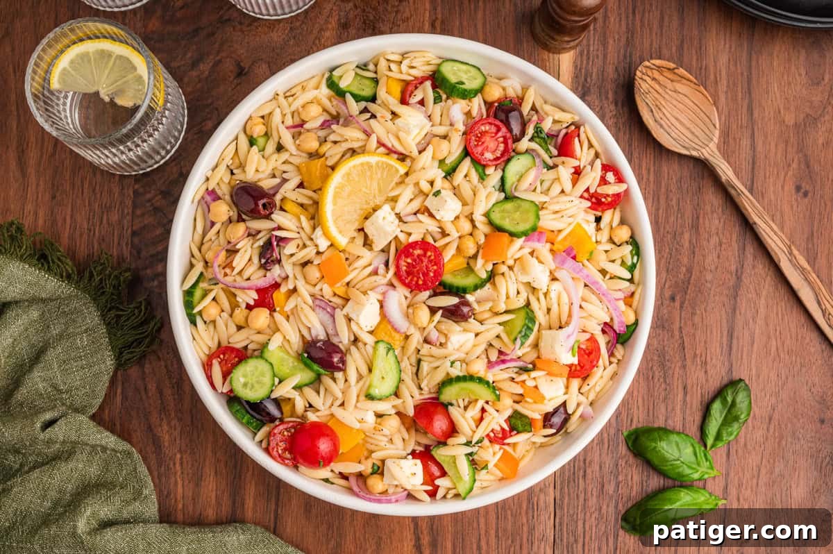Top-down view of a full bowl of orzo salad with feta, cucumbers, tomato, chickpeas, bell peppers, and olives on a wooden surface.