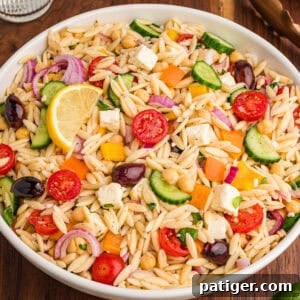 A bowl filled with orzo salad, highlighting visible ingredients such as halved cherry tomatoes, cucumber slices, red onion, chickpeas, yellow and orange bell pepper pieces, kalamata olives, cubed feta, and chopped herbs, with a lemon wedge on top.