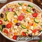 A bowl filled with orzo salad, highlighting visible ingredients such as halved cherry tomatoes, cucumber slices, red onion, chickpeas, yellow and orange bell pepper pieces, kalamata olives, cubed feta, and chopped herbs, with a lemon wedge on top.