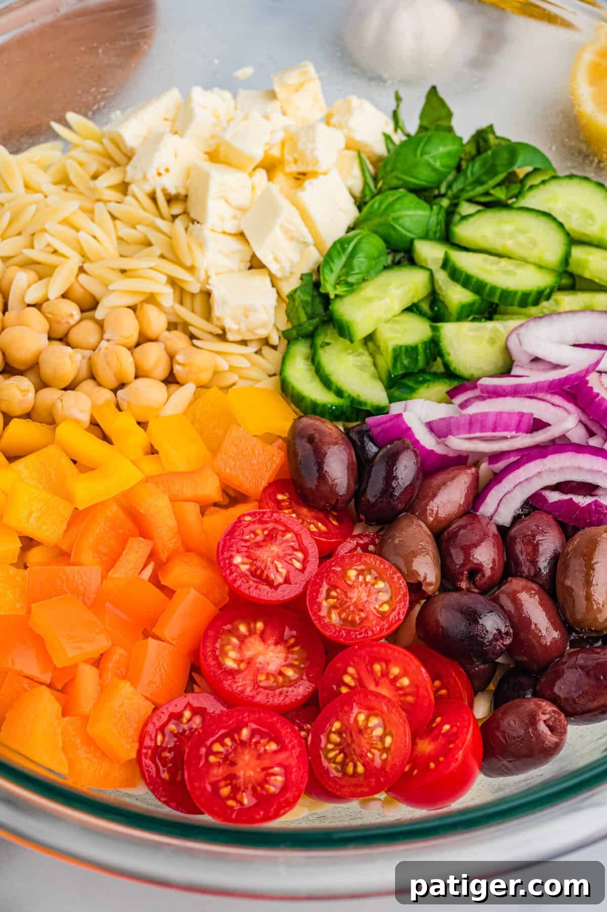 Close-up image of a glass mixing bowl with orzo salad ingredients placed in neat sections, including cherry tomatoes, kalamata olives, orange and yellow bell peppers, chickpeas, feta, basil, cucumber, and red onion.
