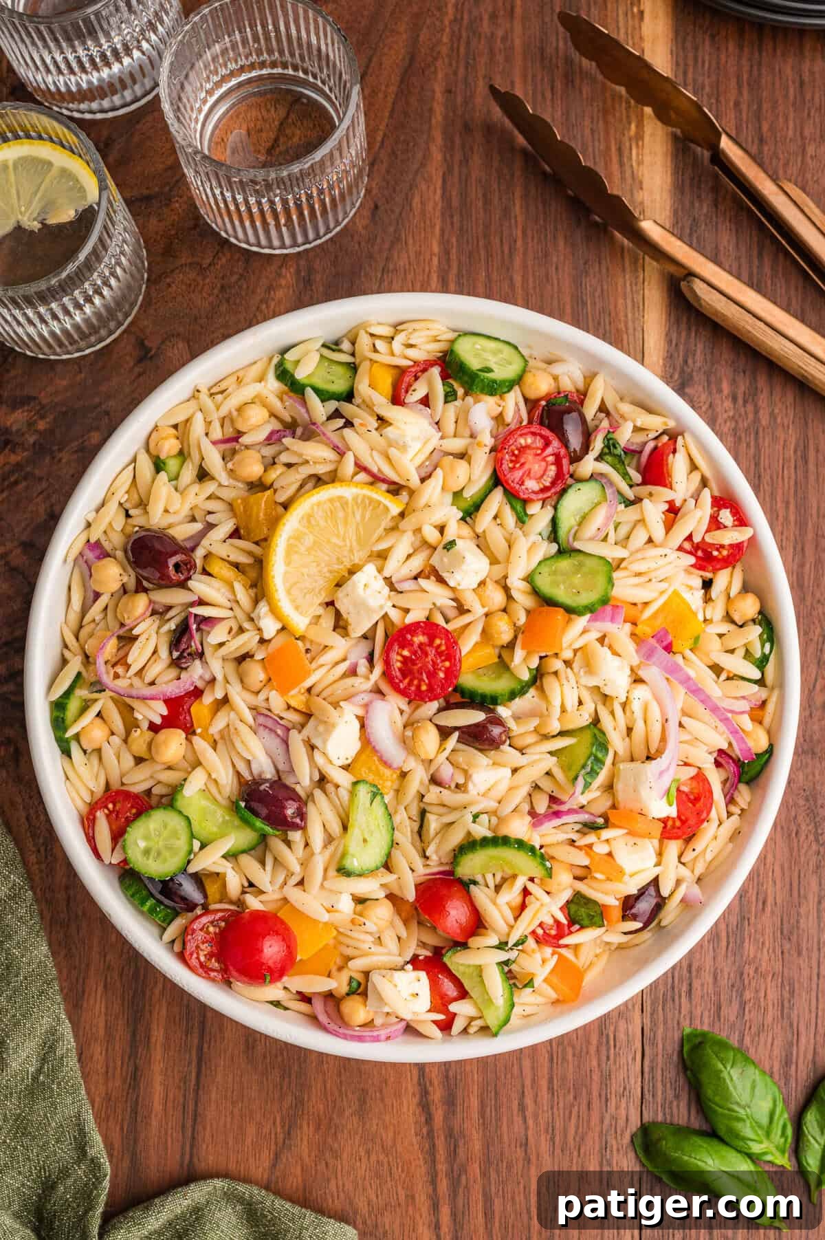 Overhead view of a large white bowl filled with orzo salad containing sliced cucumbers, cherry tomatoes, bell peppers, red onions, chickpeas, kalamata olives, feta cheese, and lemon garnish, placed on a wooden table with clear glasses of lemon water and bronze utensils in the background.