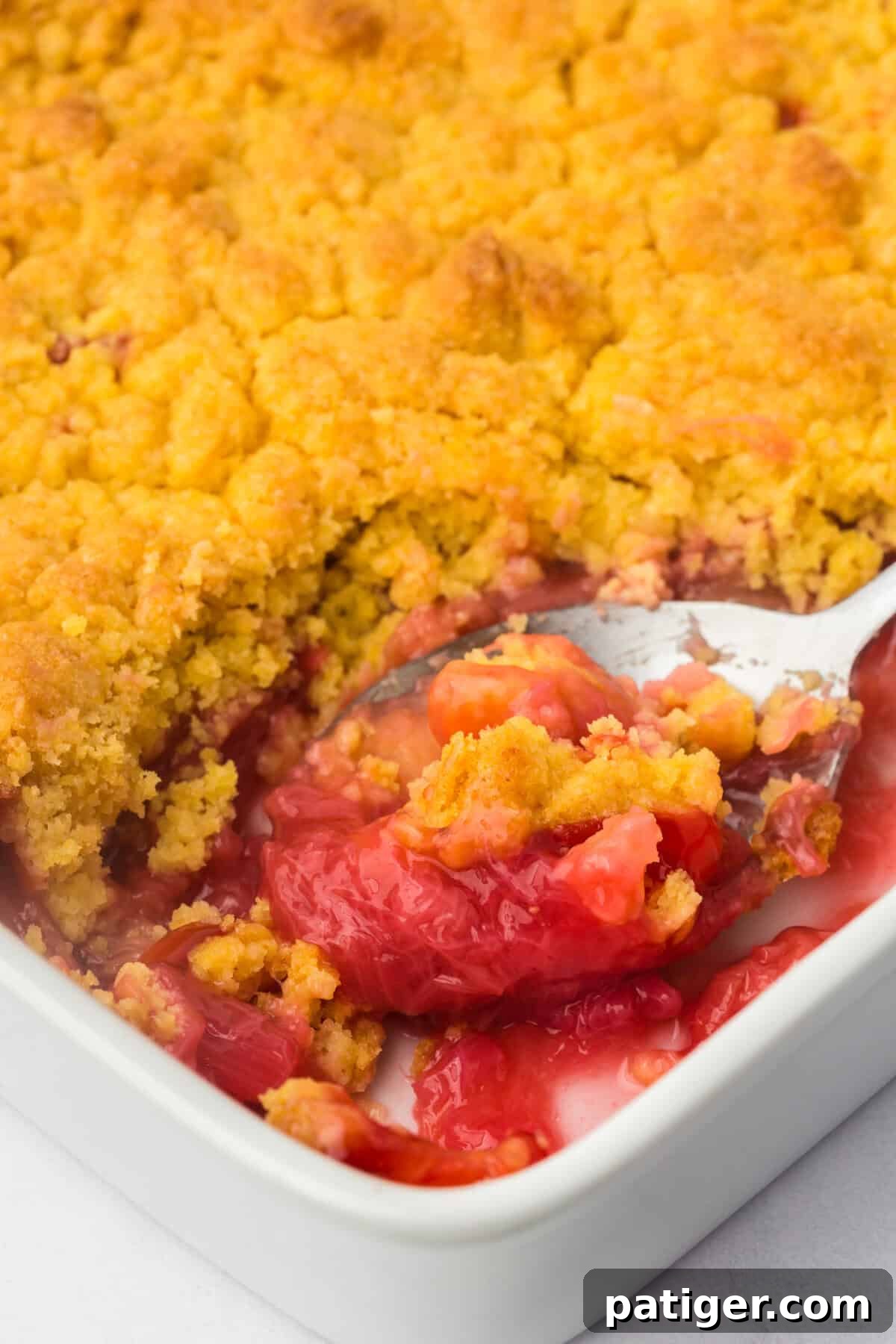 A spoon lifting a serving of strawberry rhubarb dump cake from a white baking dish. The golden, crunchy crust gives way to the bright pink-red, bubbly fruit filling underneath, showcasing the perfect texture contrast.