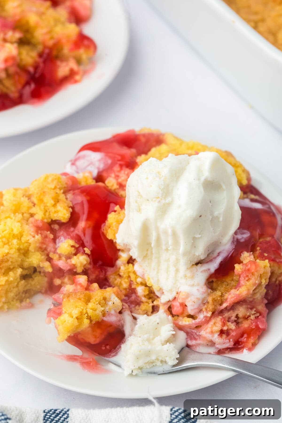 A close-up shot of a plated serving of strawberry rhubarb dump cake, with a scoop of melting vanilla ice cream cascading over its golden-brown, crumbly topping and vibrant red fruit layers. This emphasizes the luscious texture and visual appeal.