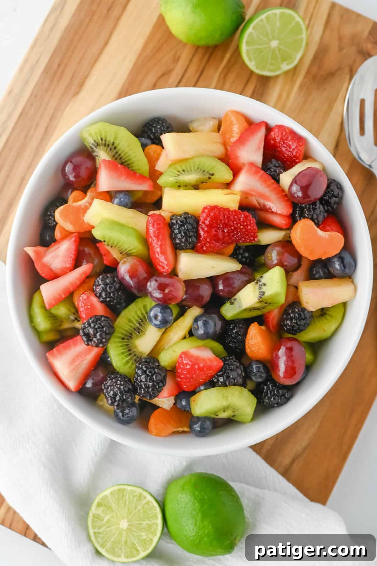Colorful fruit salad with honey lime dressing in a white bowl. Features sliced strawberries, kiwi, pineapple, red grapes, blackberries, blueberries, and clementine slices. The bowl sits on a wooden board with halved limes nearby, emphasizing freshness.
