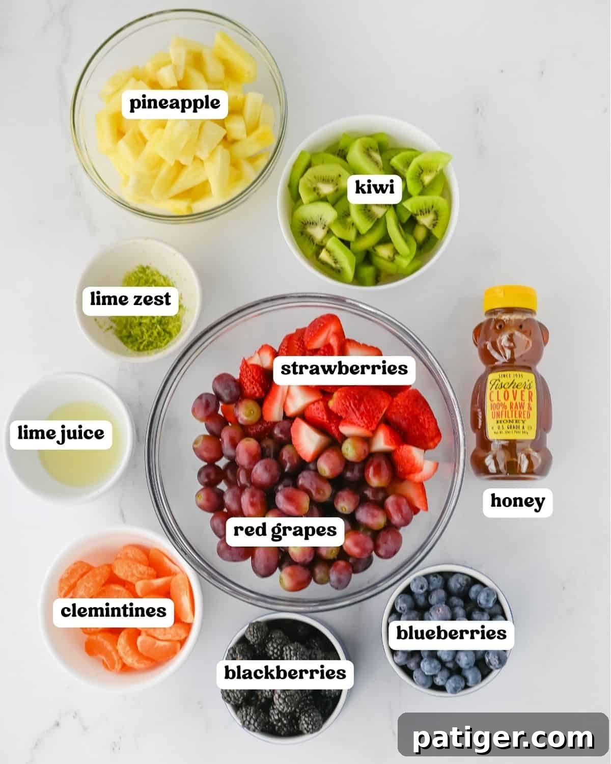 Close-up of fruit salad ingredients laid out, including a whole pineapple, sliced kiwi, fresh strawberries, clusters of red grapes, handfuls of blueberries and blackberries, clementines, a lime for zest and juice, and a jar of honey.