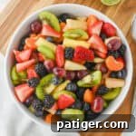 Fruit salad with honey lime dressing in a white bowl with sliced strawberries, kiwi, pineapple, grapes, blackberries, blueberries, and clementine slices.