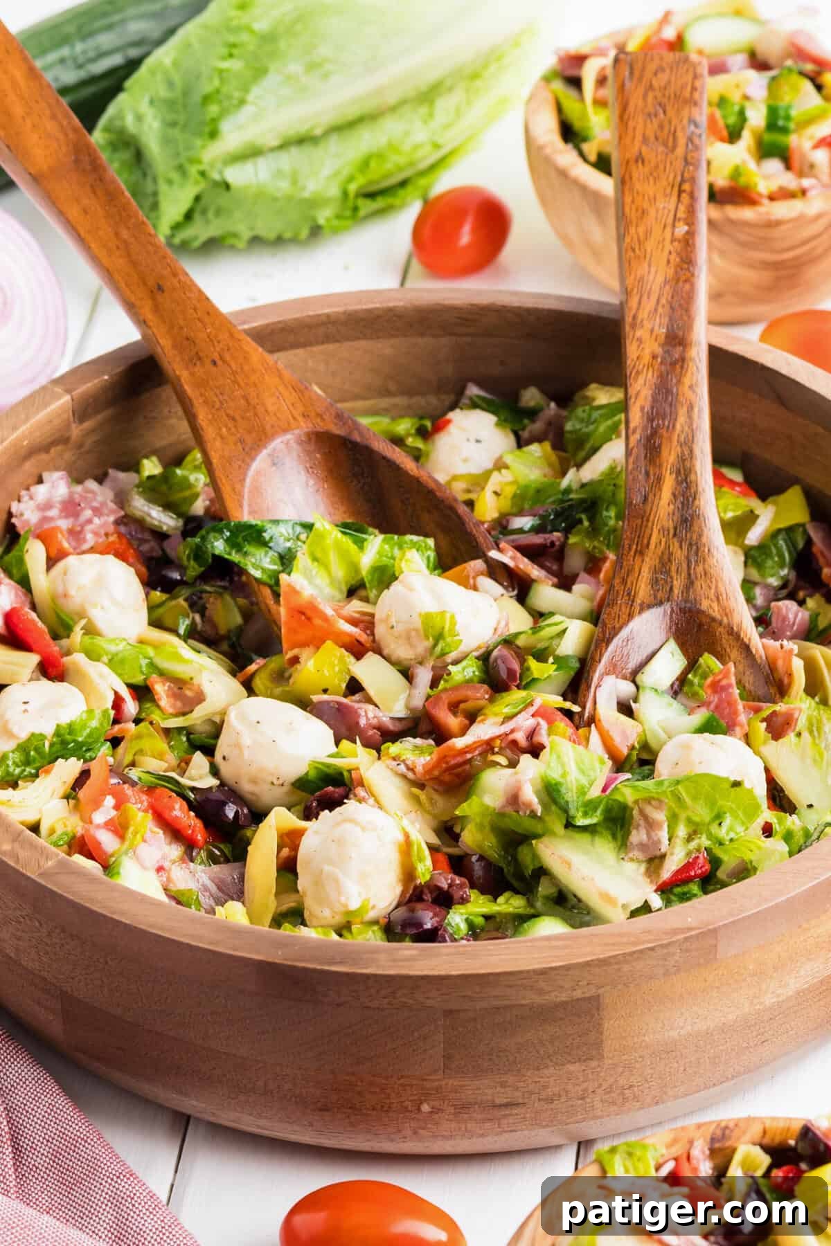 Chopped antipasto salad in a wooden bowl with  wooden serving utensils, surrounded by ingredients like romaine, cherry tomatoes, and red onion.