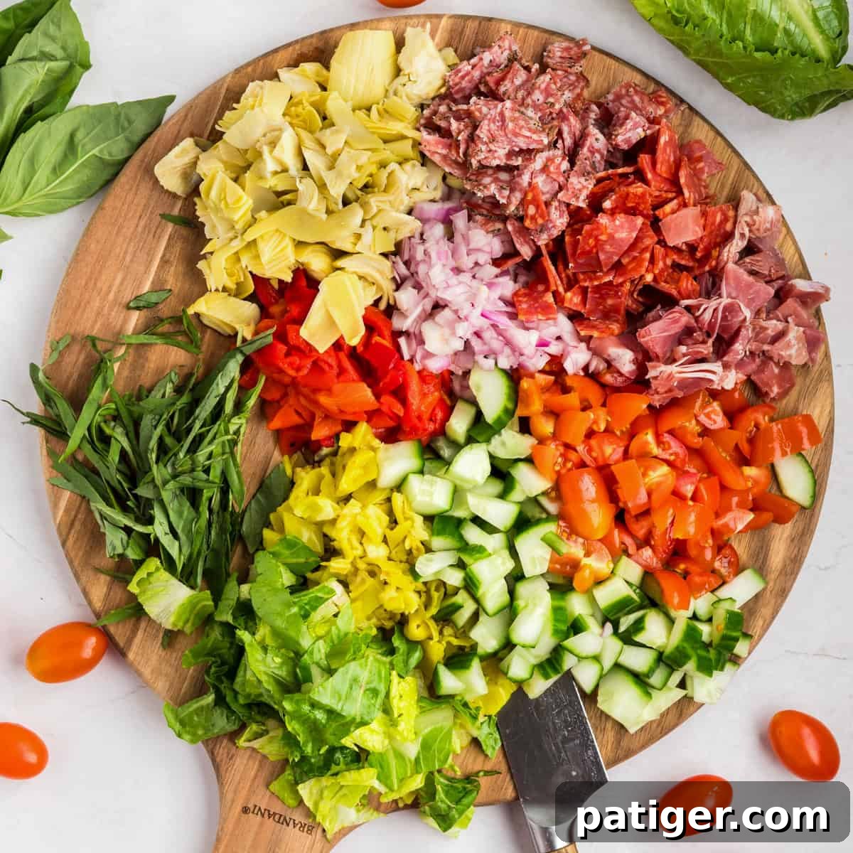Round wooden cutting board with neatly separated piles of chopped ingredients including basil, romaine, cucumbers, tomatoes, red peppers, onions, meats, artichokes, and pepperoncini.