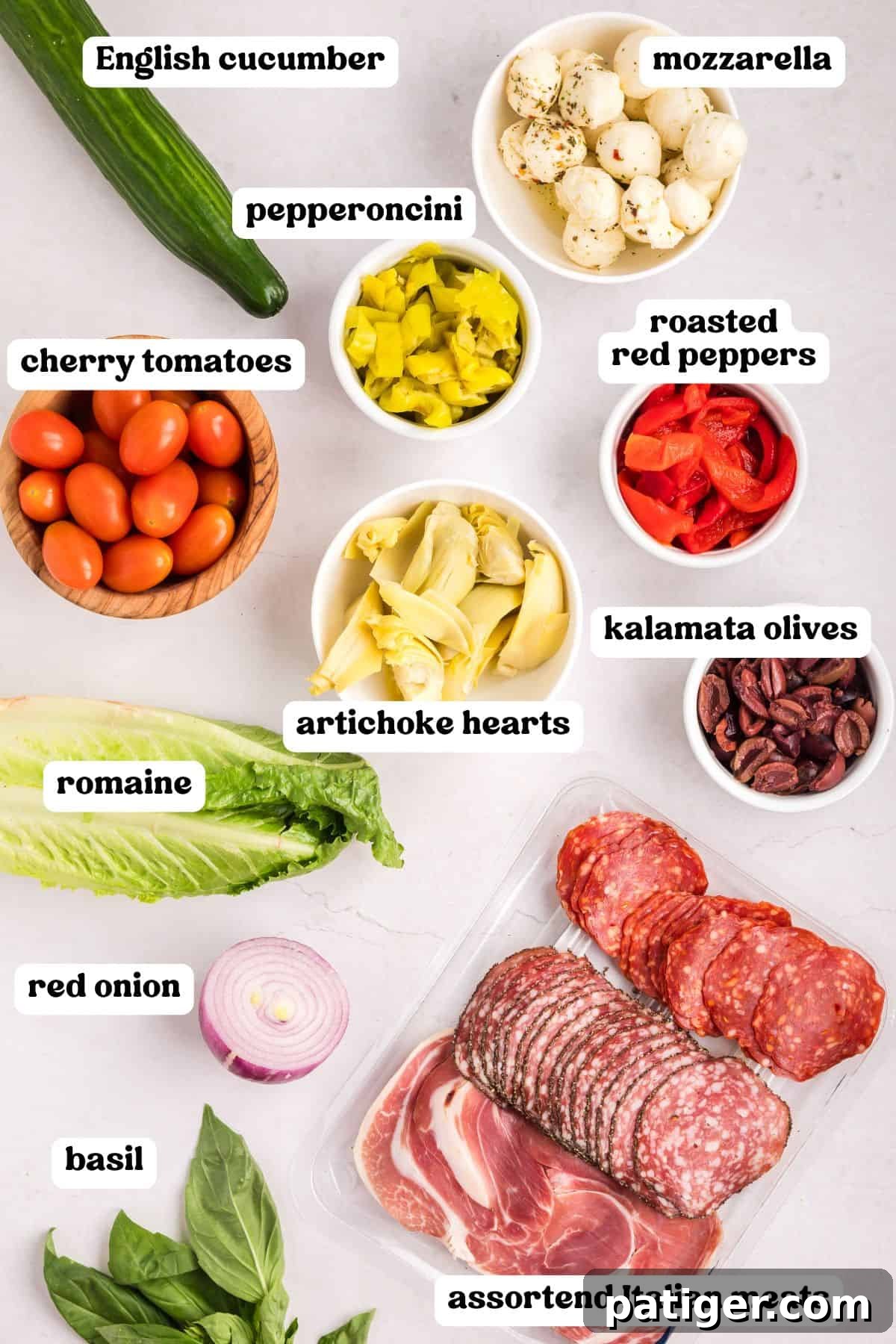 Flat lay of antipasto salad ingredients, including labeled mozzarella, pepperoncini, cherry tomatoes, roasted red peppers, kalamata olives, artichoke hearts, cucumber, red onion, romaine, basil, and assorted Italian meats.