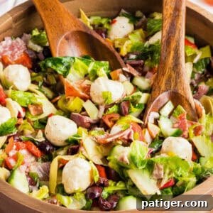 Chopped antipasto salad with mozzarella, meats, lettuce, and vegetables, in a wooden bowl with wooden serving utensils.
