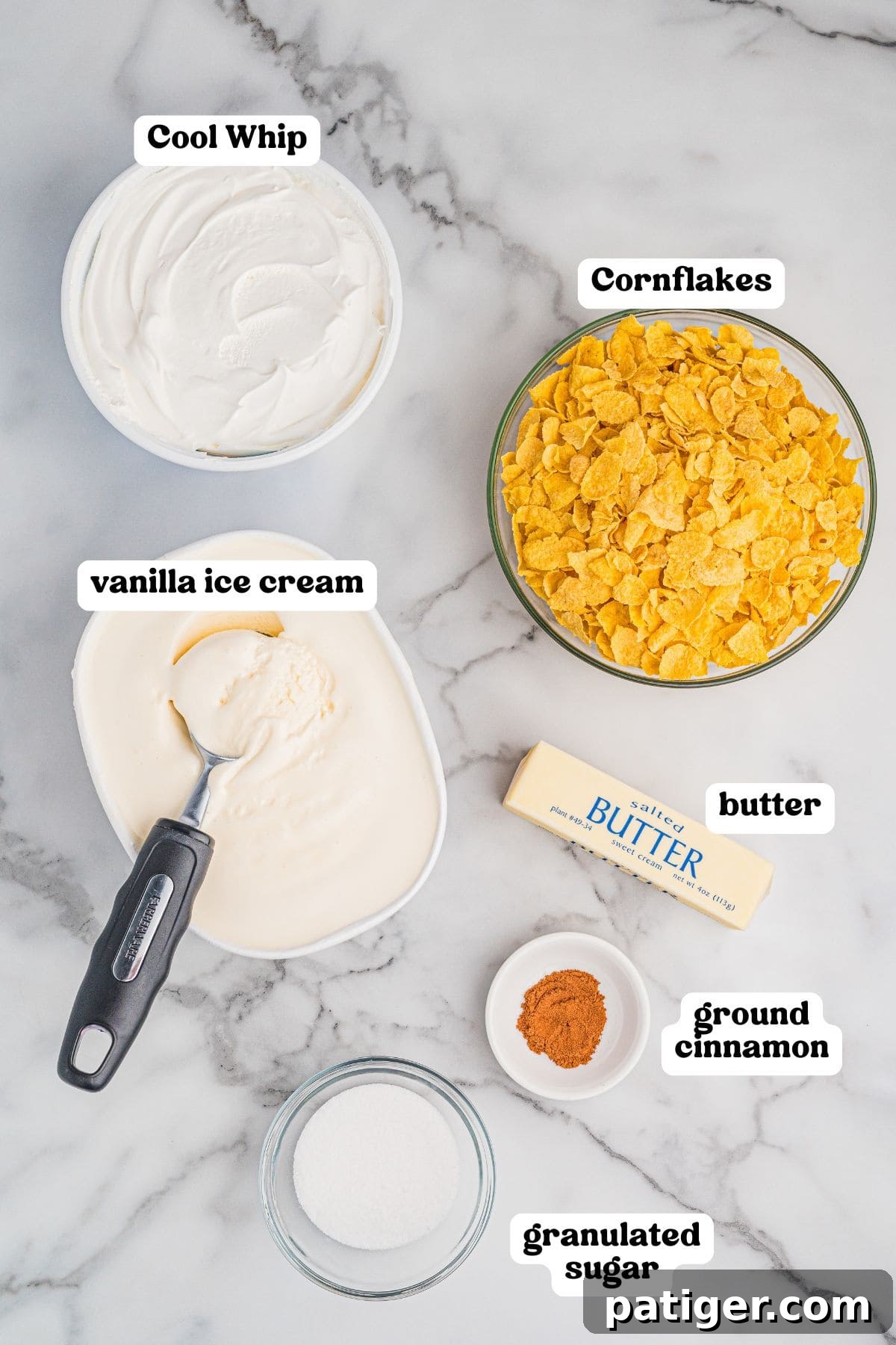 Golden Fried Ice Cream 4 Ingredients for fried ice cream arranged on a marble surface, each labeled. Includes a tub of Cool Whip, a bowl of cornflakes, a container of vanilla ice cream with a scoop, a stick of butter, a small dish of ground cinnamon, and a small bowl of granulated sugar.