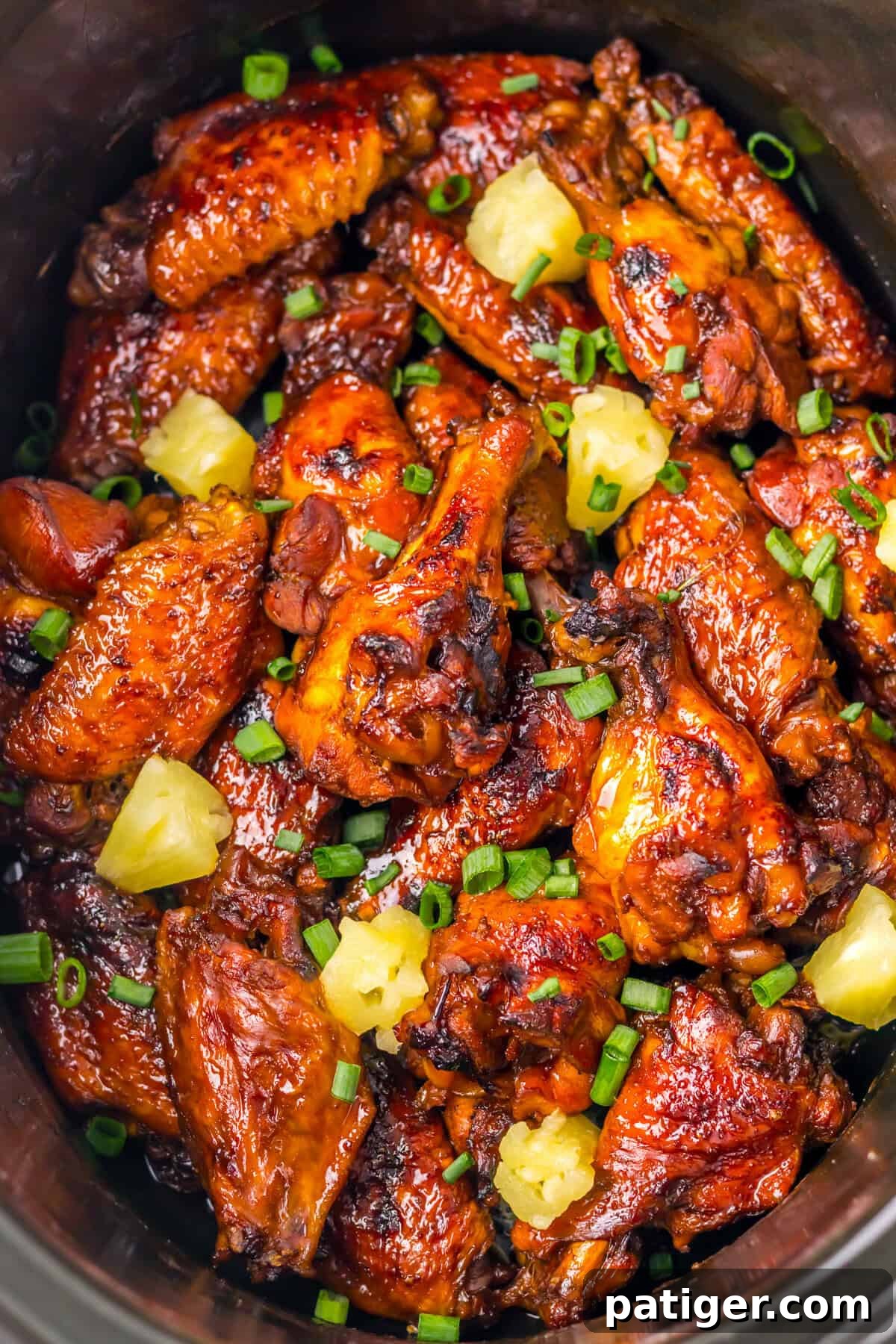 A crockpot packed with glazed Hawaiian wings. The chicken wings are topped with chunks of pineapple and chopped scallions, ready to be served.